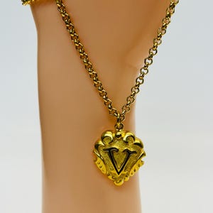 May include: A gold-colored necklace with a heart-shaped pendant. The pendant features the letter "V" in black within a decorative frame. The chain is a delicate link style, and the overall design is ornate and vintage-inspired.