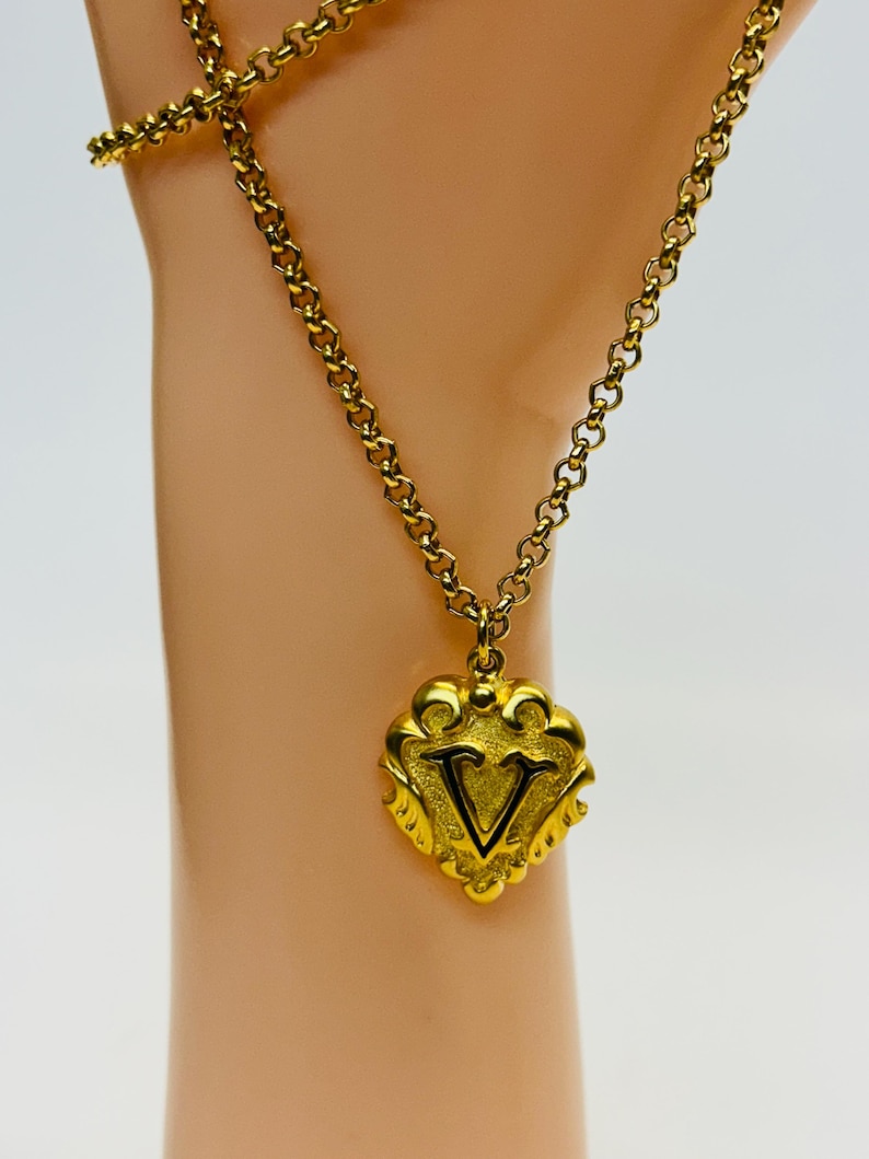 May include: A gold-colored necklace with a heart-shaped pendant. The pendant features the letter "V" in black within a decorative frame. The chain is a delicate link style, and the overall design is ornate and vintage-inspired.