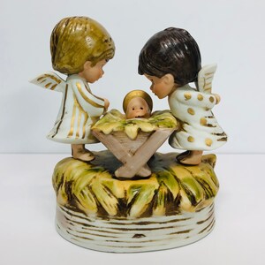 May include: A porcelain Christmas nativity scene with two angels looking down at the baby Jesus in a manger. The angels are wearing white robes with gold accents and have brown and blonde hair. The manger is made of wood and has a straw base.