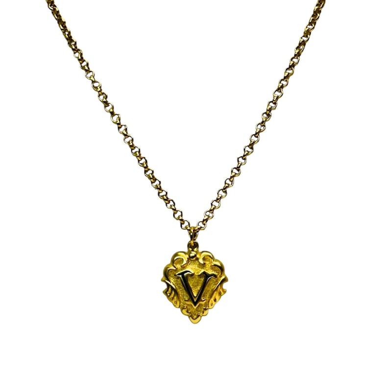 May include: A gold-colored necklace with a pendant featuring the letter "V". The pendant has an ornate, heart-shaped design. The chain is a delicate link style, and the background is white.