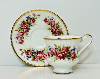 May include: A white teacup and saucer with a gold rim and floral pattern. The pattern features pink and purple roses with green leaves.