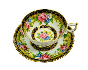 Peut inclure: Une tasse à thé et une soucoupe blanches avec un bord noir et doré. La tasse et la soucoupe sont décorées d'un motif floral dans des tons de rose, bleu et jaune.