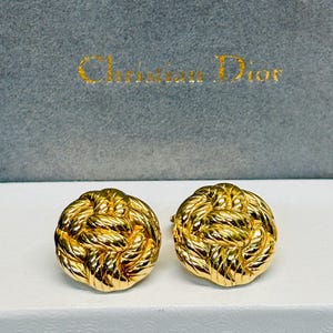 May include: A pair of gold-toned circular earrings featuring a textured, twisted rope knot design. The earrings are displayed in front of a grey box with the text Christian Dior printed in gold lettering on the front surface of the box.