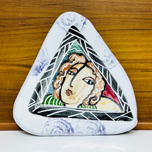 May include: A white ceramic triangular plate with a painted design of a woman's face with brown hair and a green and black patterned background.