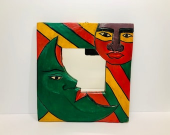 May include: A wooden mirror frame with a colorful abstract design. The frame features a green crescent moon with a face and a red and yellow striped sun with a face.