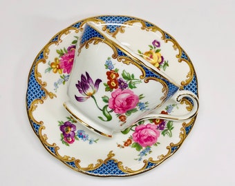 May include: A white teacup and saucer with a blue and gold floral pattern. The cup has a gold rim and a handle. The saucer has a scalloped edge.