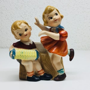 Peut inclure: Un pot de fleurs en céramique vintage représentant deux figurines d'enfants. Le garçon joue de l'accordéon et la fille danse. Le pot est peint dans des tons de brun, jaune, rouge, bleu et blanc.