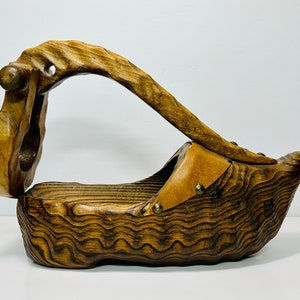 May include: A wooden fruit bowl shaped like a shoe with a leather handle. The bowl is dark brown with a wood grain pattern. The handle is attached to the bowl with metal rivets.