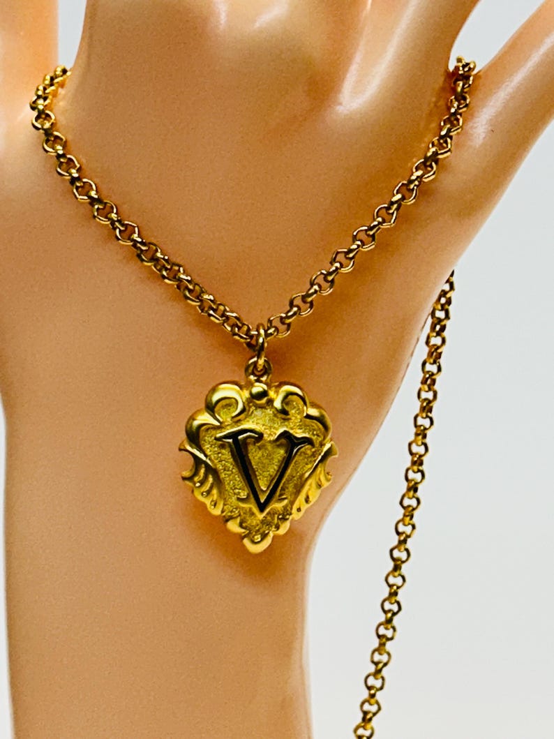 May include: A gold-colored necklace with a pendant featuring the letter "V". The pendant has an ornate, decorative design. The chain is a classic link style. The necklace is displayed against a neutral background.