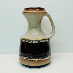 May include: A brown and beige ceramic vase with a wide mouth and a handle. The vase has a brown band with a textured edge around the middle.