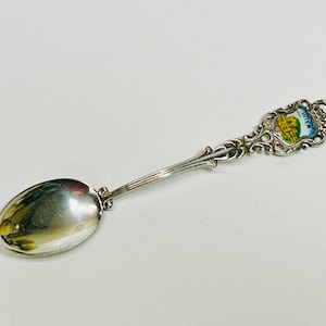 May include: A silver spoon with an ornate handle and a small, colorful emblem. The emblem features a building and the word "WIEN". The spoon has a reflective, oval bowl.