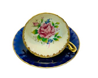 Peut inclure: Une tasse à thé et une soucoupe vintage avec un motif floral. La tasse est blanche avec un bord doré et une rose rose avec des feuilles vertes et des fleurs bleues. La soucoupe est bleu foncé avec un bord doré.