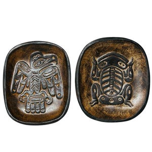 May include: Two dark brown wooden bowls with rounded square edges. The left bowl features a carved eagle design, while the right bowl displays a carved frog design. Both bowls have a distressed, textured finish with deep black accents in the carvings.