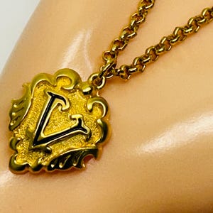 May include: A gold-toned necklace featuring a pendant with a stylized letter "V" in black enamel. The pendant has an ornate, scrolled border. The chain is a matching gold color and has a textured, linked design. The necklace is displayed on a light-colored surface.