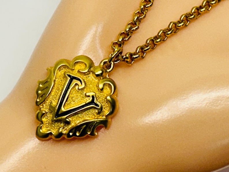May include: A gold-toned necklace featuring a pendant with a stylized letter "V" in black enamel. The pendant has an ornate, scrolled border. The chain is a matching gold color and has a textured, linked design. The necklace is displayed on a light-colored surface.