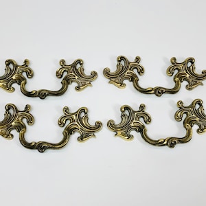 May include: Six ornate brass cabinet pulls with a decorative, swirling design. The pulls have a traditional style and are suitable for furniture restoration or crafting projects.