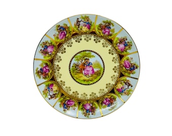 May include: A white porcelain plate with a gold rim and a delicate floral design. The center of the plate features a romantic scene with two figures in a garden setting. The plate is decorated with a repeating pattern of gold and pink flowers.