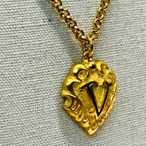 May include: Gold-toned necklace with a pendant featuring the letter "V". The pendant has an ornate, heart-shaped design. The chain is a delicate link style, and the necklace is displayed against a neutral background.