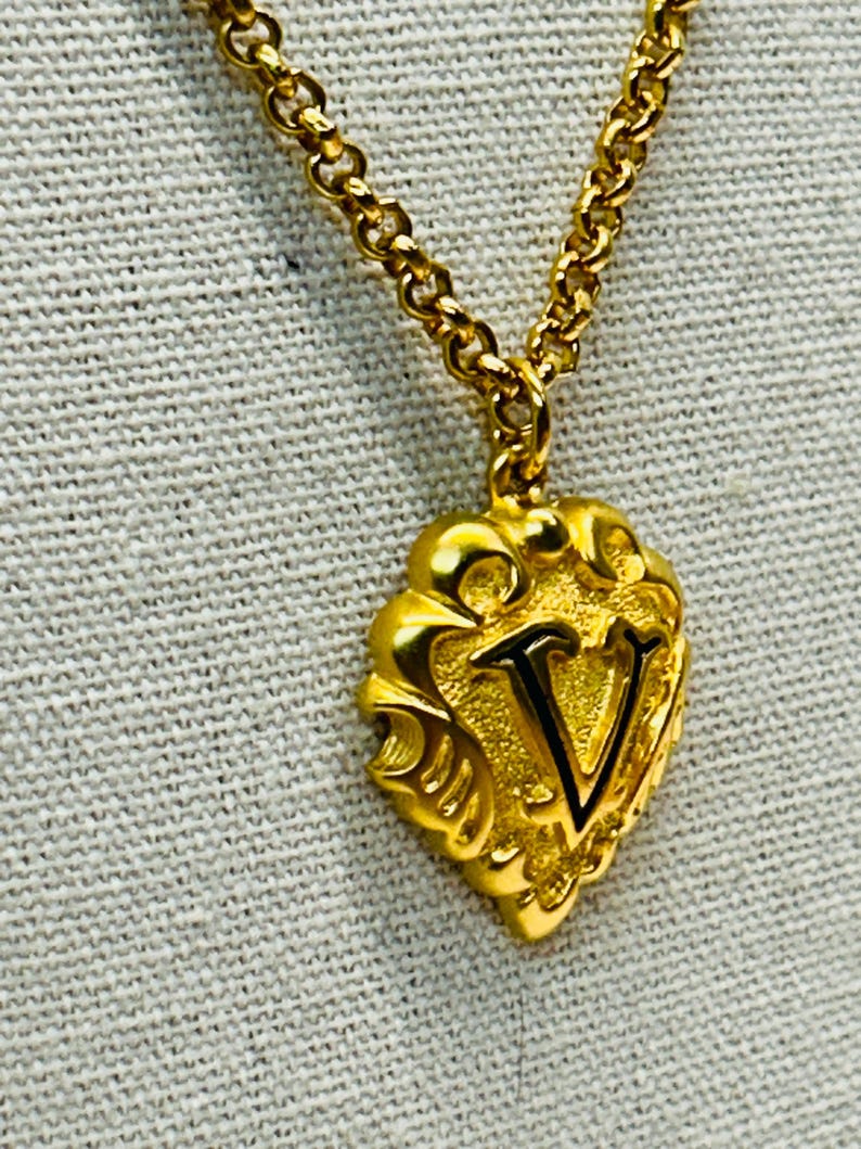 May include: Gold-toned necklace with a pendant featuring the letter "V". The pendant has an ornate, heart-shaped design. The chain is a delicate link style, and the necklace is displayed against a neutral background.