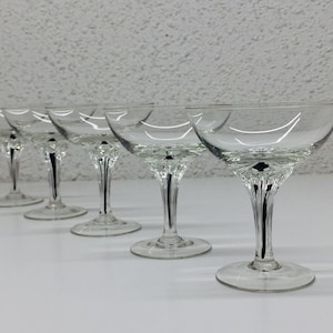 May include: Five clear glass champagne coupes with a fluted stem and a wide bowl. The glasses are arranged in a row on a white surface.