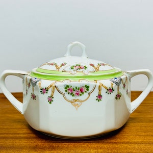 May include: White porcelain sugar bowl with a lid, featuring a floral design with pink and green roses, gold accents, and a green band around the lid.