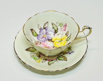 Peut inclure: Une tasse à thé et une soucoupe blanches avec un motif floral de pensées violettes, jaunes et roses. L'ensemble est bordé d'une garniture dorée.