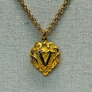 May include: Gold-toned necklace with a pendant featuring the letter "V". The pendant has an ornate, shield-like design with a black outline. The chain is a delicate, linked style, and the background is a textured, light gray fabric.