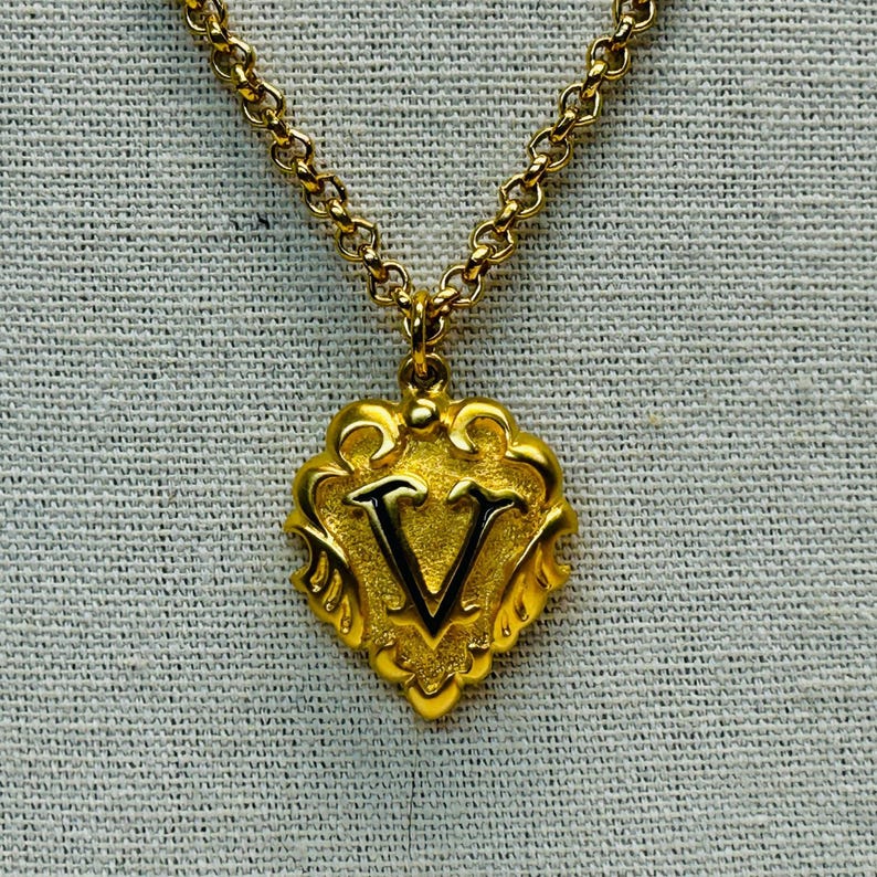 May include: Gold-toned necklace with a pendant featuring the letter "V". The pendant has an ornate, shield-like design with a black outline. The chain is a delicate, linked style, and the background is a textured, light gray fabric.
