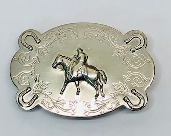 May include: Silver belt buckle featuring an intricate design of a cowboy riding a horse and four horseshoes. The buckle has a vintage appearance.
