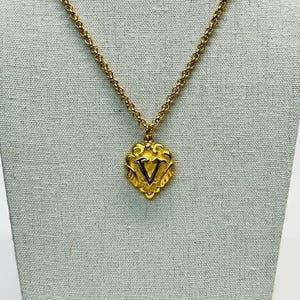 May include: Gold-toned necklace with a pendant featuring the letter "V". The pendant is heart-shaped with ornate detailing. The chain is a delicate, linked design. The necklace is displayed against a neutral backdrop.