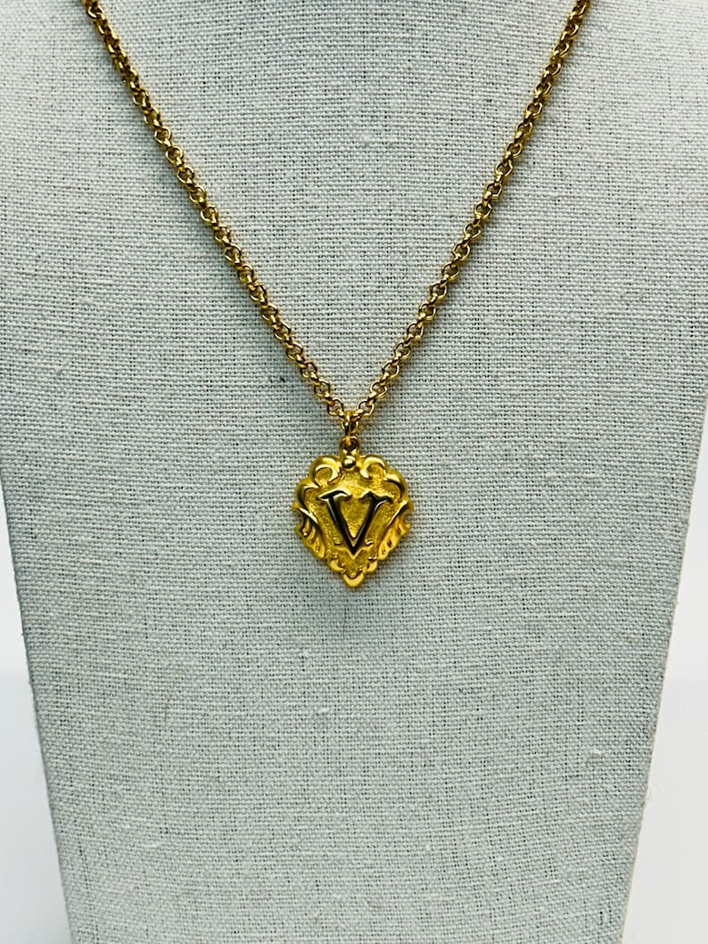 May include: Gold-toned necklace with a pendant featuring the letter "V". The pendant is heart-shaped with ornate detailing. The chain is a delicate, linked design. The necklace is displayed against a neutral backdrop.