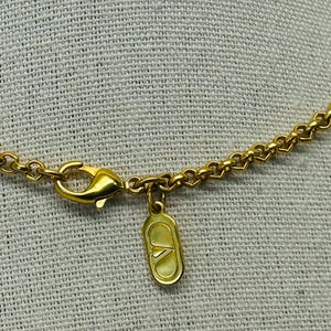 May include: Close-up of a gold-colored necklace with a lobster clasp and a small, rectangular charm. The chain has a heart-shaped design. The charm has a logo on it. The necklace is on a light-colored, textured background.