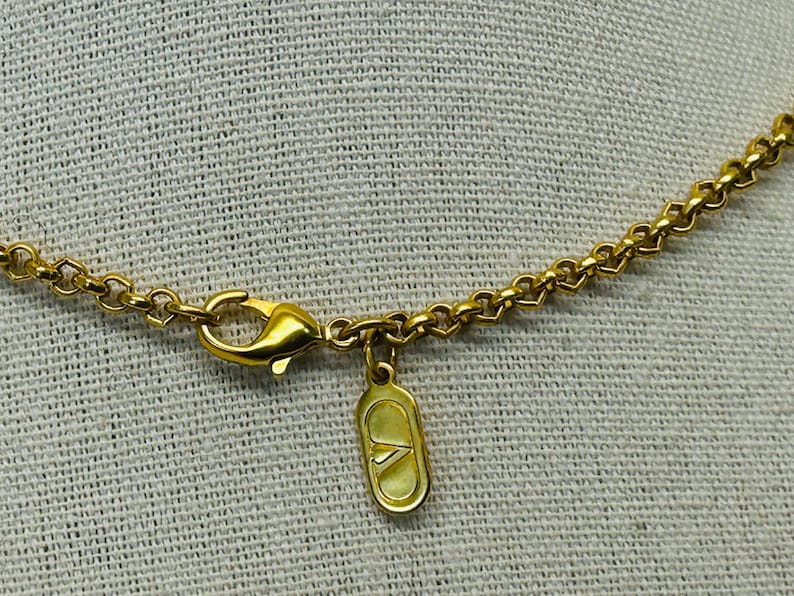 May include: Close-up of a gold-colored necklace with a lobster clasp and a small, rectangular charm. The chain has a heart-shaped design. The charm has a logo on it. The necklace is on a light-colored, textured background.