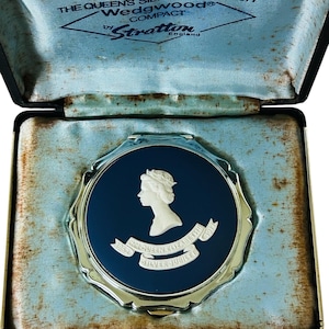 Könnte beinhalten: Ein Vintage-Kompakt in einem schwarzen Etui. Das Kompakt hat ein blaues Gesicht mit einem weißen Profil und dem Text "THE QUEEN'S SILVER JUBILEE 1977". Das Etui ist mit blauem Satin gefüttert und enthält den Text "Wedgwood Compact by Stratton England".
