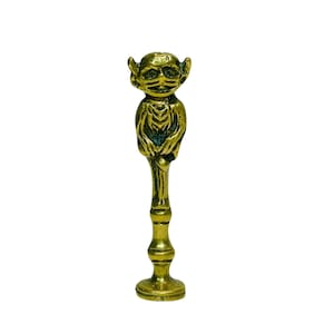 May include: A brass-coloured figurine of a goblin-like creature. The figurine has a detailed face with pointed ears and is holding its hands together. The figure is mounted on a decorative stem and a round base. The overall design is ornate.