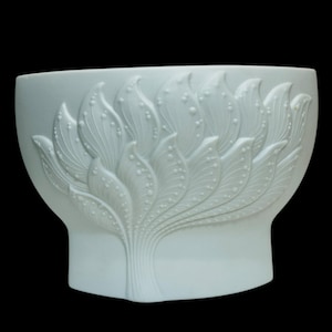 May include: A white ceramic planter with a detailed, raised floral design. The design features stylized leaves and small, round accents. The planter has a wide, open top and a slightly flared base, set against a black background.