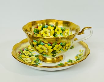 May include: A gold teacup and saucer set with a floral pattern of yellow primroses. The cup and saucer have a scalloped edge and are decorated with gold trim.