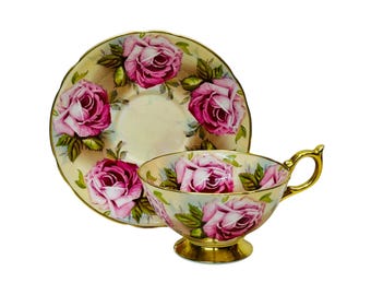 May include: A vintage teacup and saucer set. The cup and saucer feature pink rose and green leaf designs on a cream base. The cup has a gold handle and base, and the saucer has a scalloped edge with gold trim.