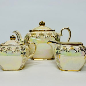 May include: A white tea set with gold accents. The teapot, sugar bowl, and creamer are all decorated with a delicate floral pattern. The set is made of porcelain and has a glossy finish.