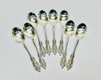 May include: Eight silver spoons with ornate handles arranged in a fan shape on a white background.