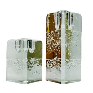 Peut inclure: Deux bougeoirs en verre transparent. L'un est de forme carrée et court, l'autre est de forme rectangulaire et plus grand. Les deux ont un aspect texturé semblable à de la glace et sont conçus pour contenir des bougies. Les bougeoirs sont transparents.