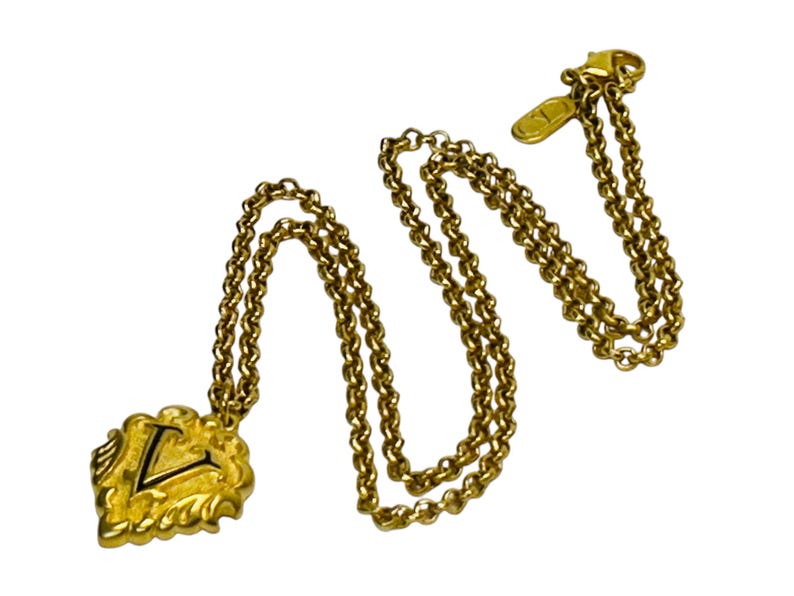 May include: Gold-toned necklace with a pendant featuring the letter "L" in black. The pendant has a decorative, ornate design. The chain is a classic link style, and the clasp is visible. The necklace is set against a white background.