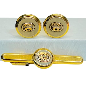 May include: A set of gold-toned metal cufflinks and a matching tie bar, each featuring a circular design with a central cream-colored inlay and the iconic interlocking G logo in gold. The accessories are presented together as a coordinated set.