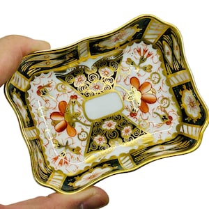 May include: A square, scalloped-edge, porcelain dish with a black, gold, and red floral pattern. The dish is decorated with a gold trim and has a white center.
