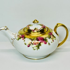 Peut inclure: Une théière en porcelaine blanche avec une bordure dorée et un motif floral composé de roses roses et blanches. La théière a un couvercle avec un bouton doré.