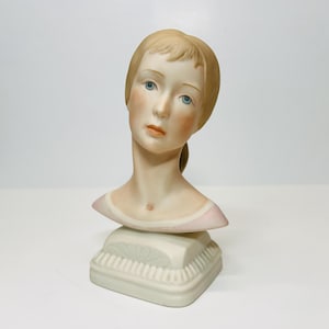 May include: A porcelain bust of a young woman with blonde hair and blue eyes. She is wearing a pink dress and has a delicate, feminine expression.