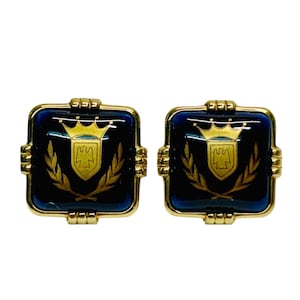 Puede incluir: Un par de gemelos cuadrados con un centro de cristal azul profundo enmarcado en metal dorado. La cara de cada gemelo muestra un emblema dorado de una corona sobre un escudo con un motivo de ave, flanqueado por dos ramas de laurel.