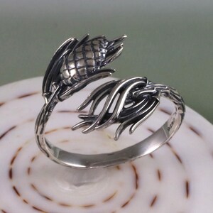May include: A silver ring with a detailed design of a pine cone and branches. The ring is open and wraps around the finger.
