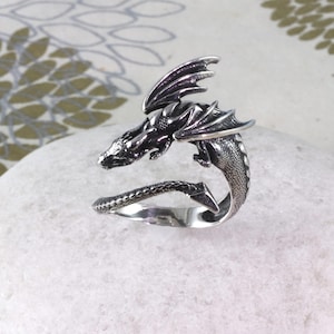 May include: A silver dragon ring with a detailed design. The dragon is curled around the ring band, with its wings spread out.