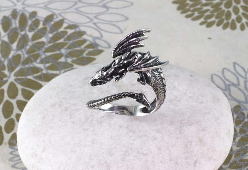 May include: A silver dragon ring with a detailed design. The dragon is curled around the ring band, with its wings spread out.